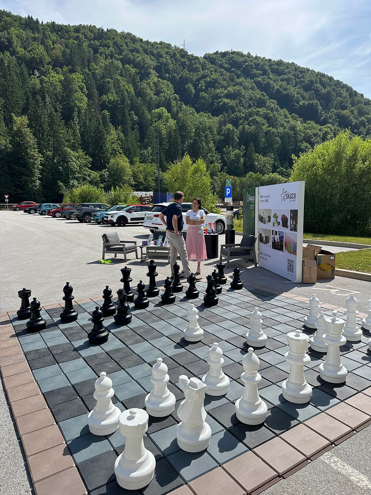 Moški in ženska se pogovarjata ob stojnicah poleg velikega zunanjega šahovskega polja z belimi in črnimi figurami