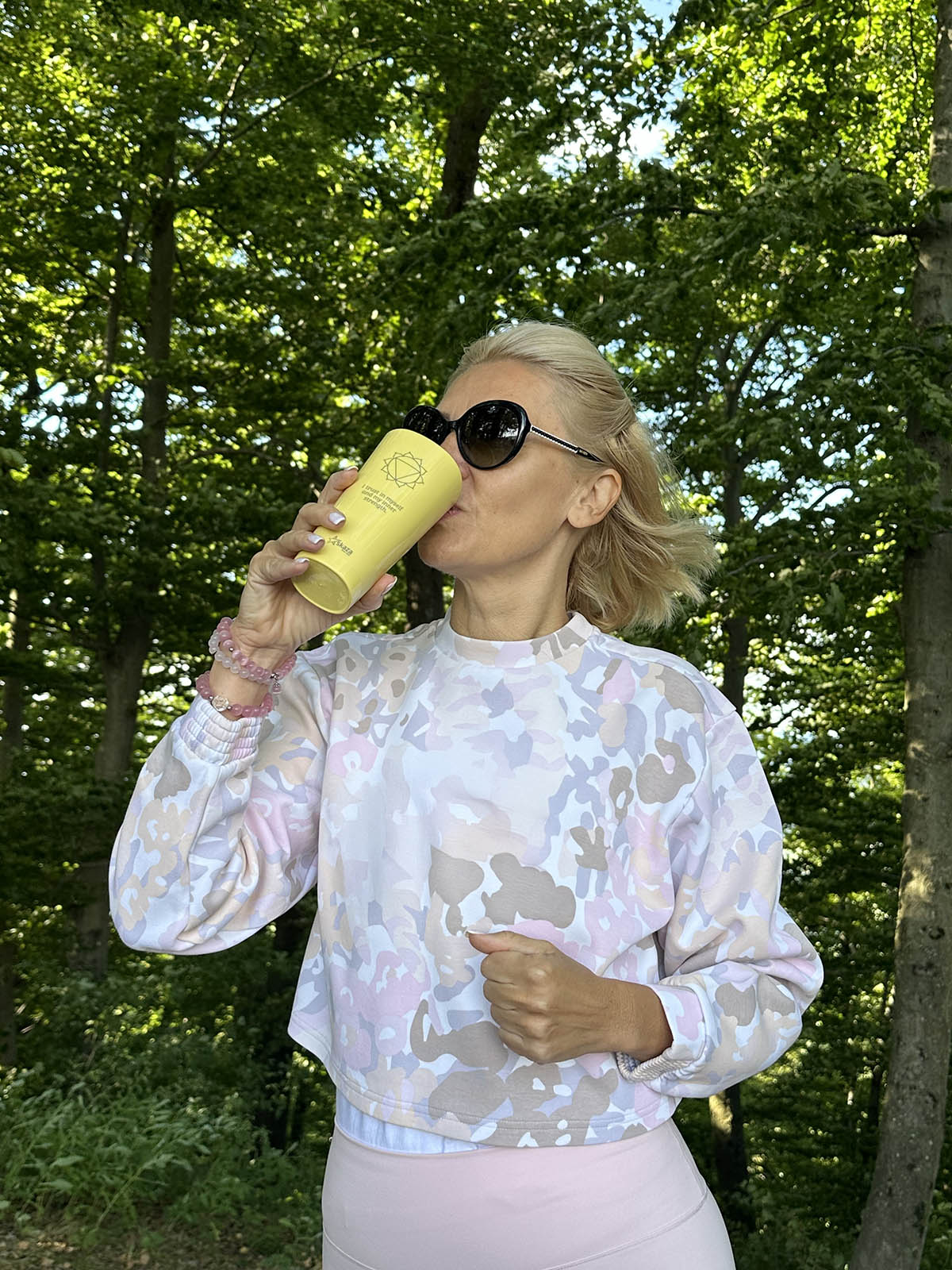 Tanja Skaza v športni opravi pije iz rumenega trajnostnega kozarca Skaza z motivom solarnega pleksusa med sprehodom po gozdu, obdana z zelenjem in jutranjo svetlobo.