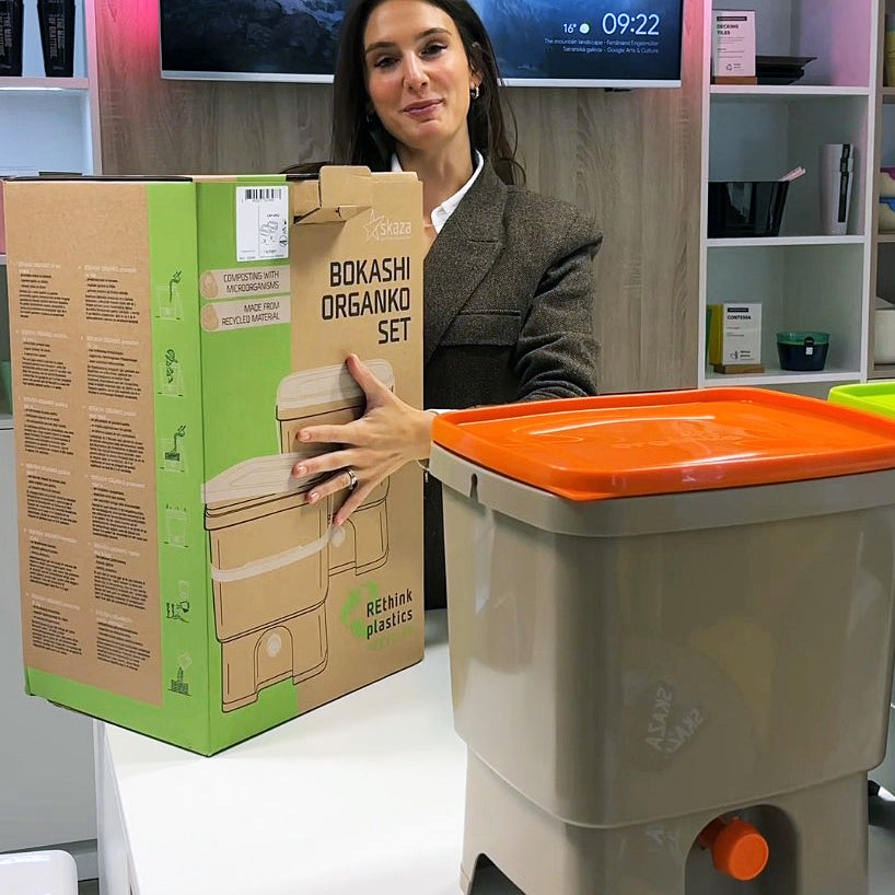 Ženska drži embalažo kompleta Bokashi Organko 1, ob njej je postavljen kompostnik v kremno-oranžni barvi.