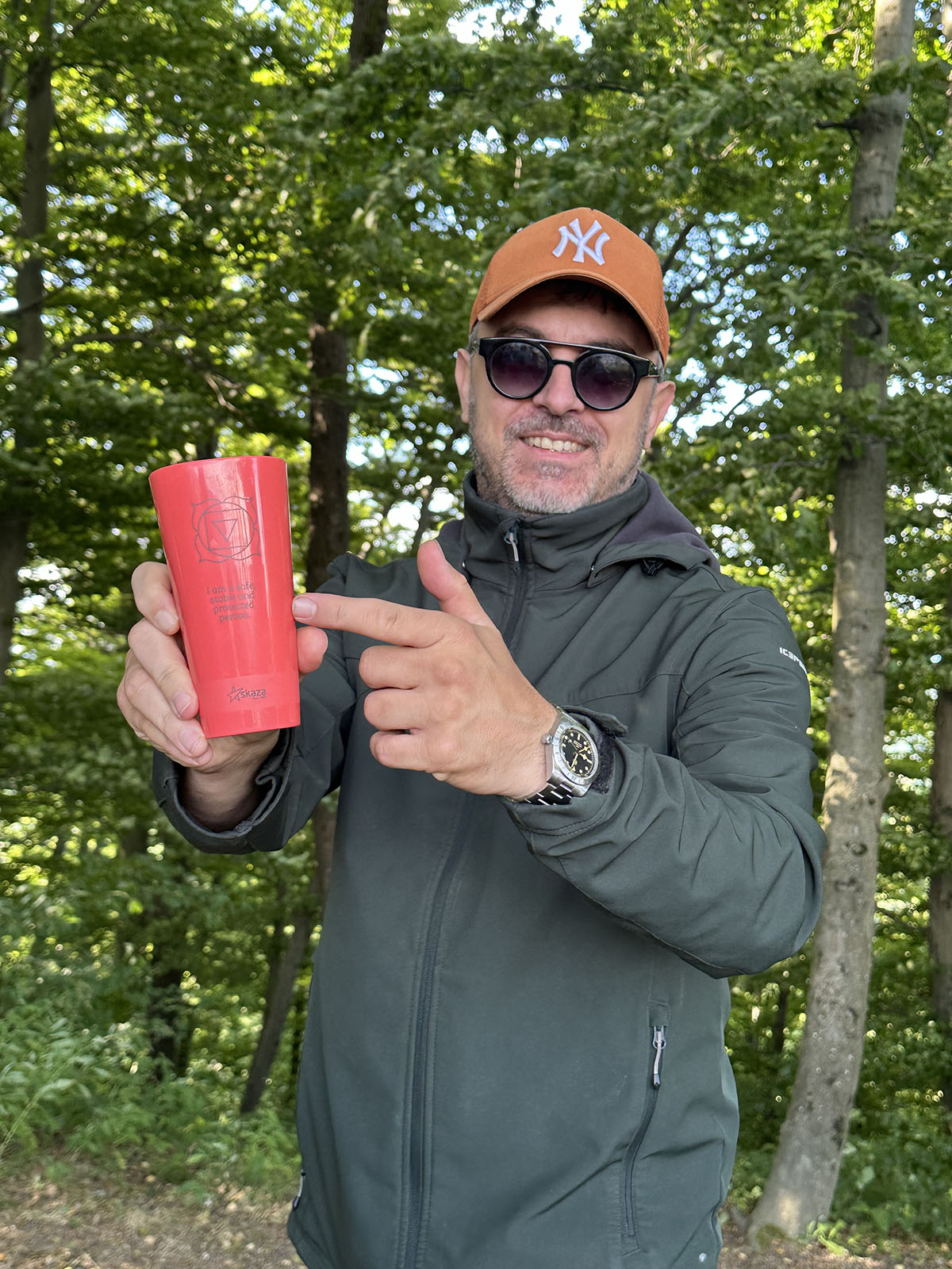 Moški z nasmehom stoji v gozdu, nosi oranžno kapo in sončna očala, ter v rokah drži rdeč trajnostni kozarec Skaza s simbolom korenske čakre in napisom: »Čutim varnost. Zaupam v življenje.«