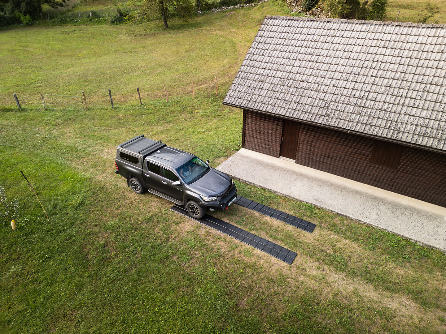 Avto parkiran na črnih pohodnih ploščah, ki tvorijo dve vzporedni poti na travnati površini ob leseni stavbi